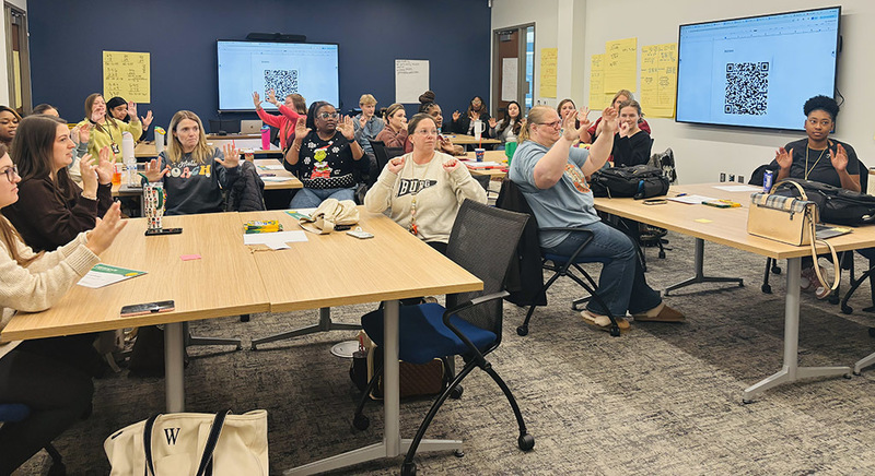Teachers seated at desks raising hands and engaging in the lesson