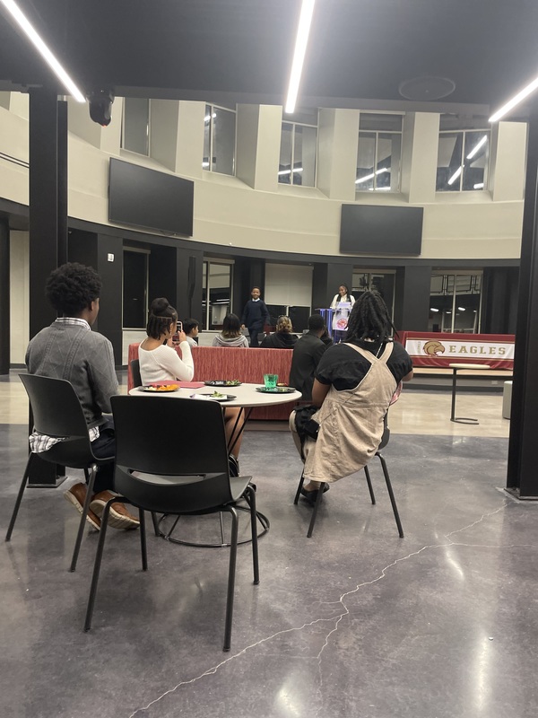 Students sitting at the table looking at Coach Haynes on the stage as she gives a speech