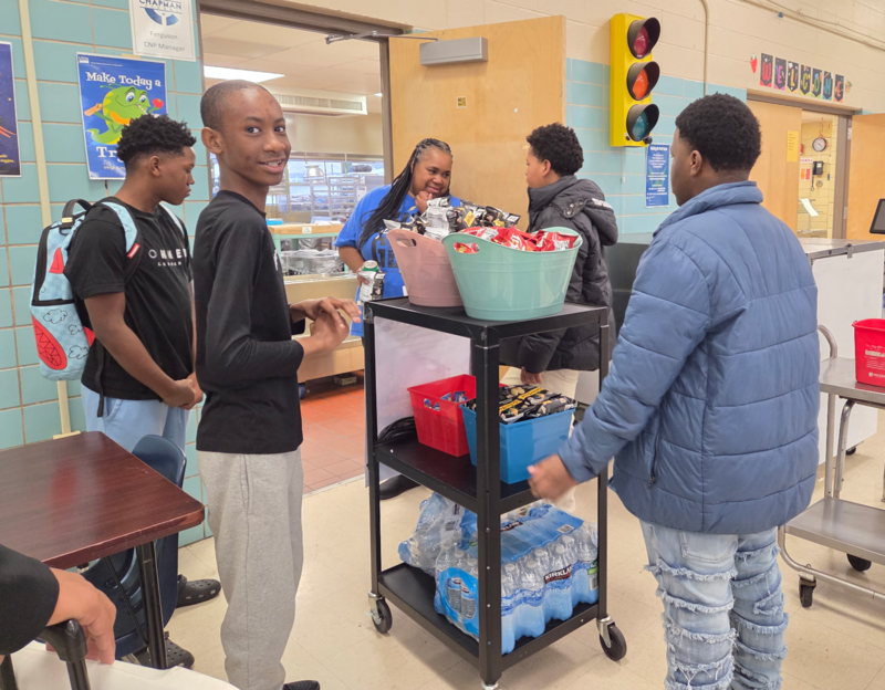 A group of students gather around the mobile snack cart in a school cafeteria, as they distribute refreshments to the staff.