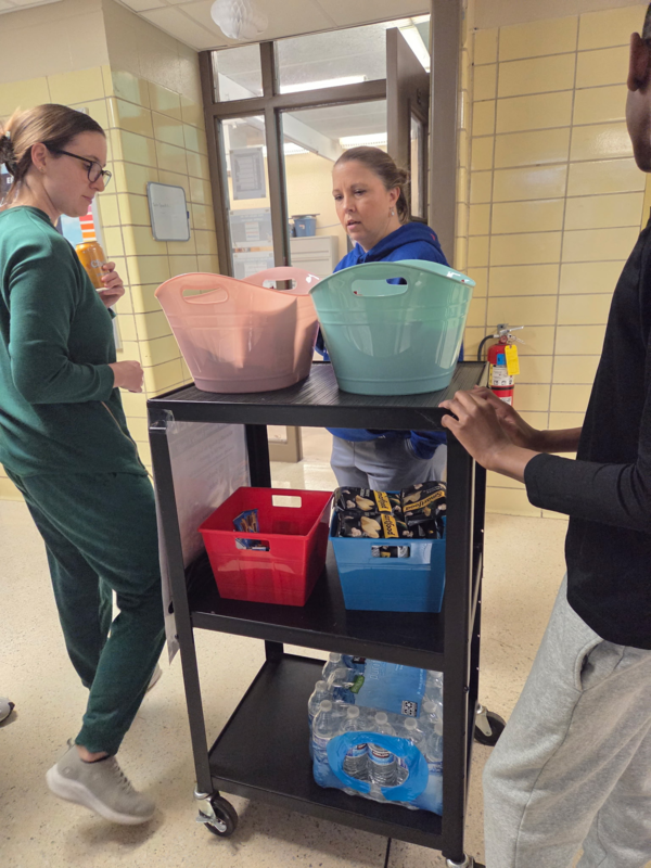 Two female staff members at Artemis Virtual Academy select items from the "Friday Fuel" cart while a student stands nearby to assist.