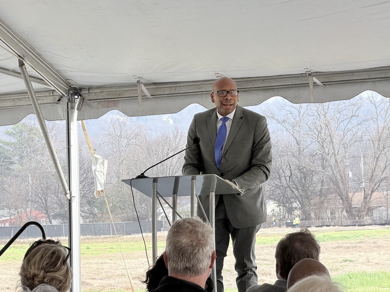 Dr. Clarence Sutton, wearing a grey suit, glasses, and a blue tie, speaks from a metal podium under a white event tent. He is looking toward an audience seated in the foreground, with an open field and trees visible in the background.