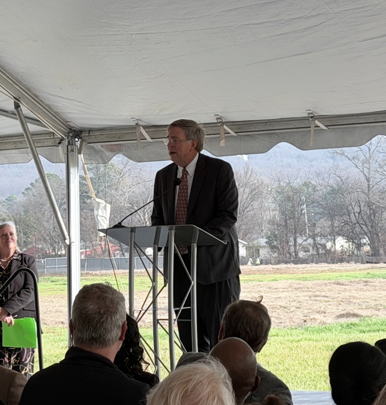 Mayor Tommy Battle, wearing a dark suit and patterned tie, speaks at a metal podium under a white event tent. A woman stands nearby holding a green folder, and an audience is seated in front of the Mayor with an open field in the background.