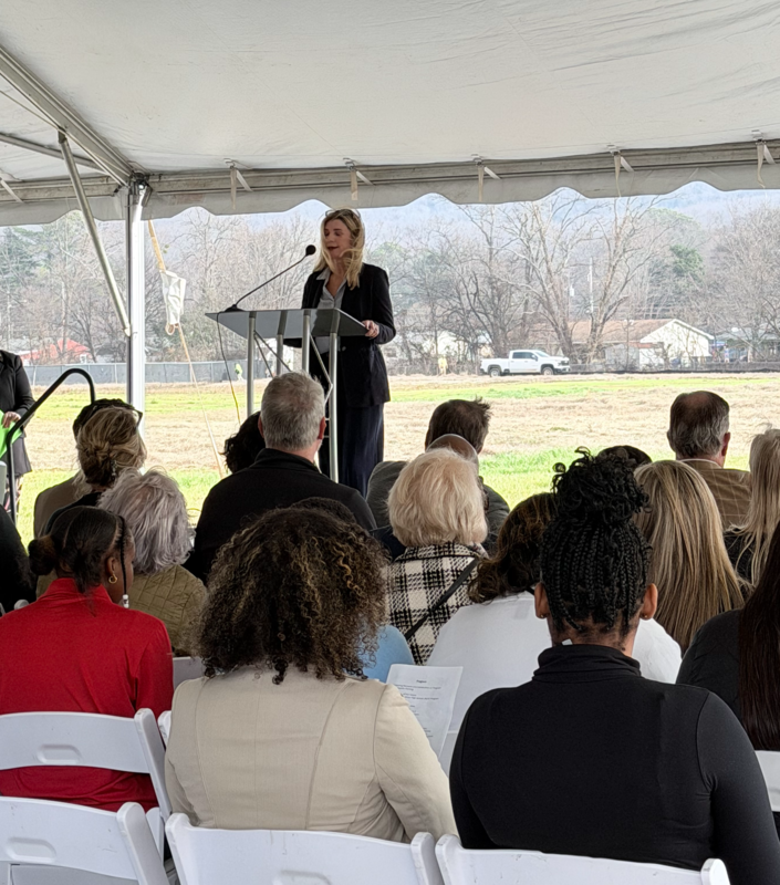 School Board Representative Holly McCarty, a woman with blonde hair in a black blazer, speaks at a metal podium under a white tent. A seated audience fills the foreground, and a white pickup truck is visible in the distant background across an open field.