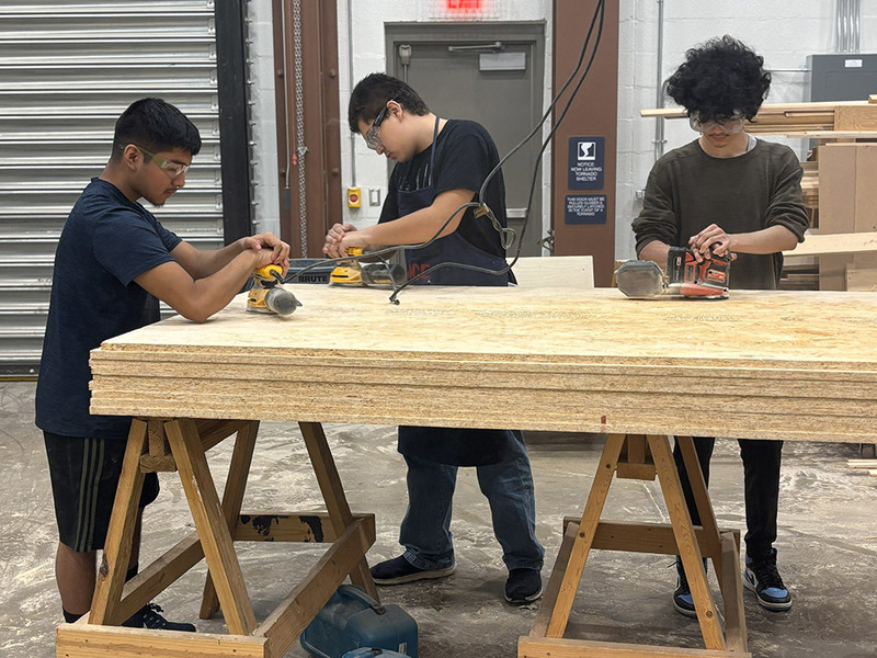 Three students sanding a piece of plywood with electric sanders