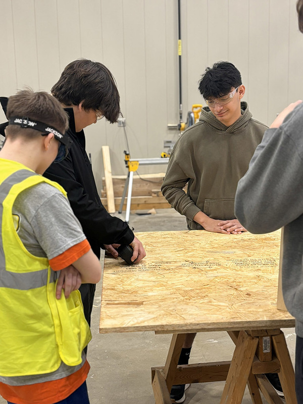 One student measures a large plank of plywood while other students observe