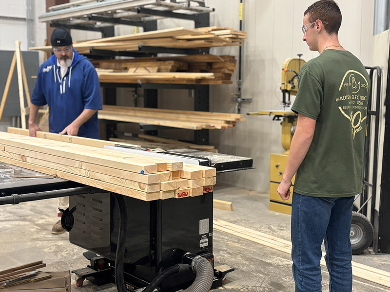 Teacher pushing board across saw while student waits to receive the other end