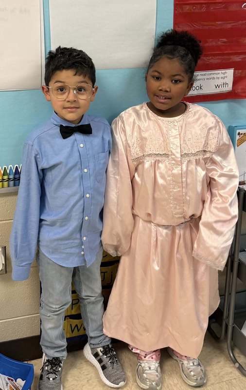 Kindergarten students that are dressed like they are 100 years old. One is in a blue shirt with a bow tie and the other students is in a pink dress.