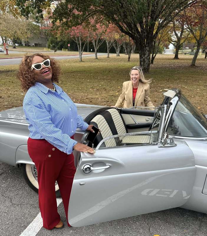 Ms. Caudle and Ms. Sutton in front of a nice car as if they are going to drive away.