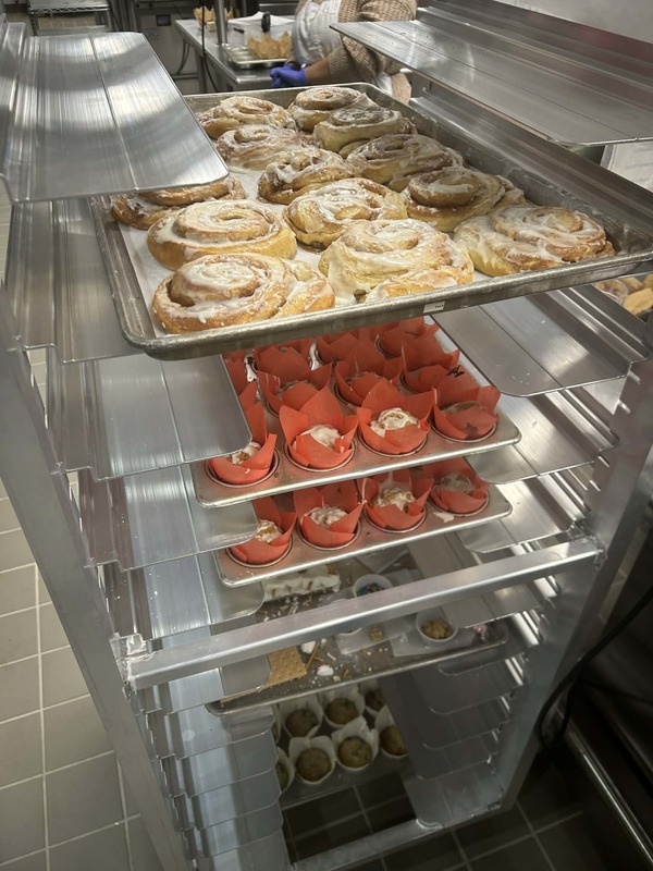 Bakery racks full of trays of cinnamon rolls and muffins in liners.