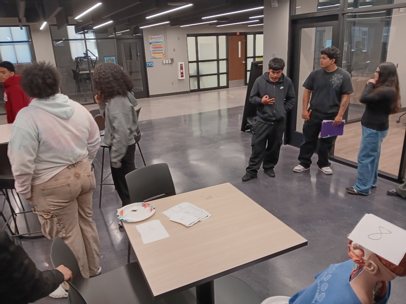 Six students standing in Innovation Center that is staged like a crime scene investigating the crime with a dummy in the chair at the table