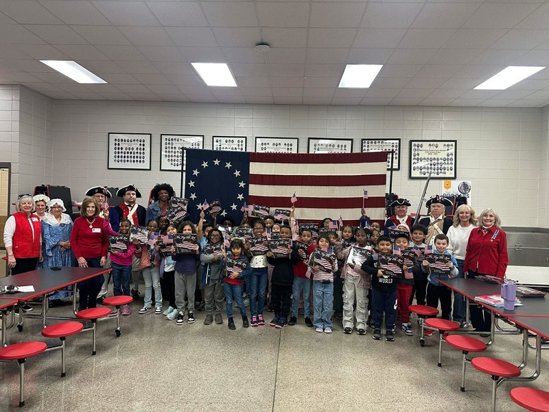 Second graders holding their books up with DAR presenters dressed up along with the American flag representing the thirteen colonies.