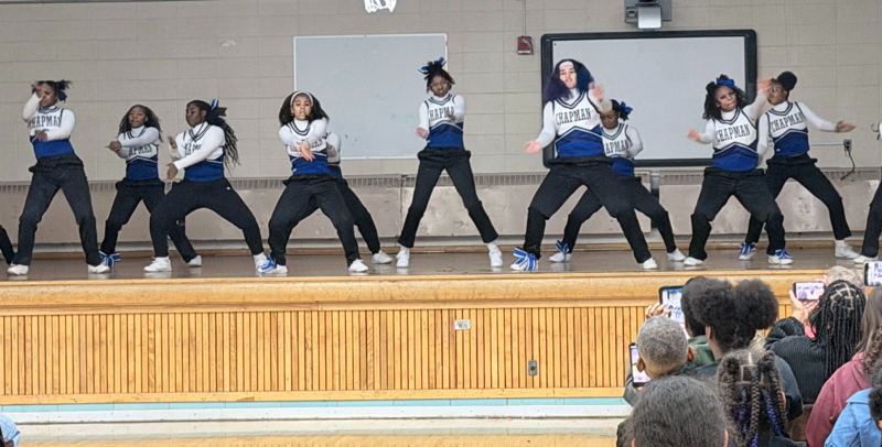 A line of Chapman Middle School cheerleaders in blue, white, and black uniforms perform a synchronized dance routine on a gym stage. They are in low, athletic stances with their arms out. 