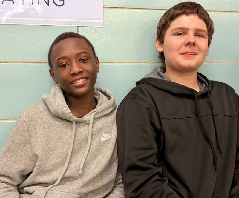 Two male students sit side-by-side against a light blue brick wall. Both students are smiling as they enjoy the talent show. 