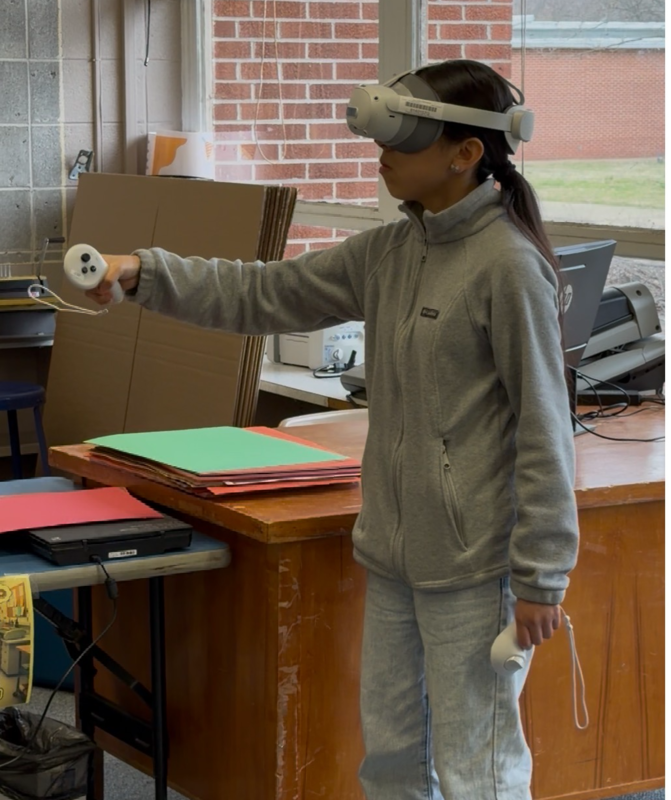 A student wearing a grey zip-up jacket and a VR headset stands near windows and a desk in the library. She is extending one controller forward while immersed in her math lesson.