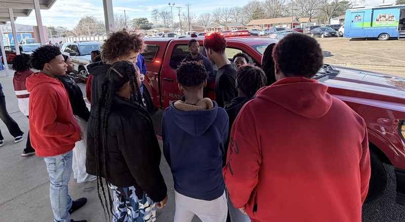 A group of students gathers outside a red Huntsville Fire Department truck while a firefighter explains the vehicle's features while discussing firefighter expectations.