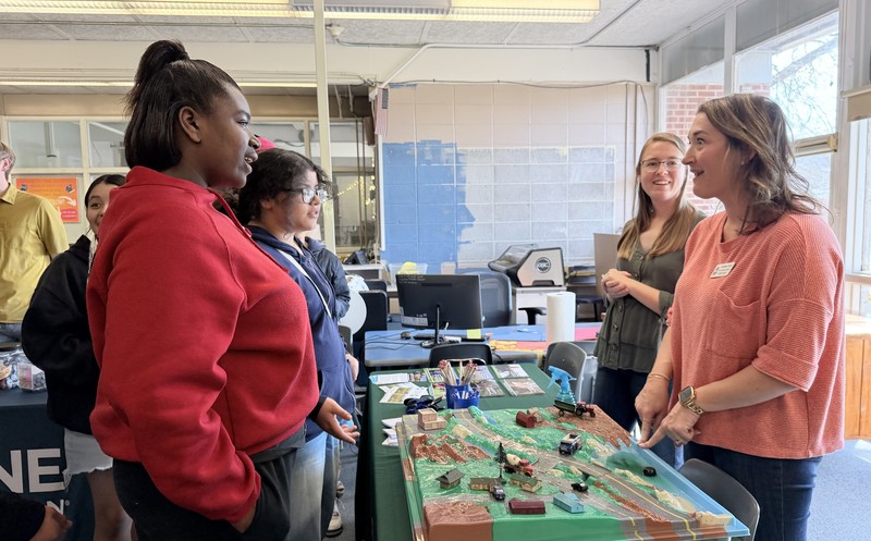 A female presenter points to a 3D environmental model of a landscape and river while students look on.