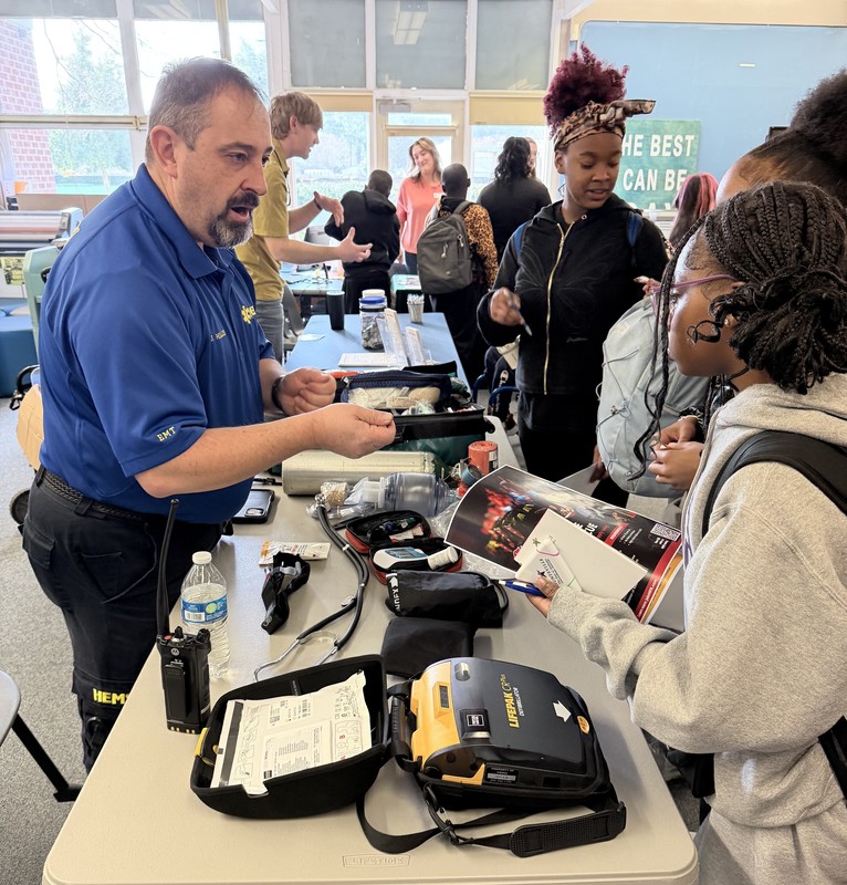 A male EMT in a blue uniform interacts with students at a table covered in medical equipment, including a stethoscope and a LifePak defibrillator.