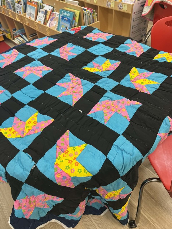 A quilt with bright blue squares, yellow and pink design in the middle, and a black outline of the squares.