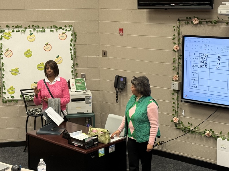2 women from AKA soroity dressed in pink and green speaking to class standing around desk with white board in the back of them