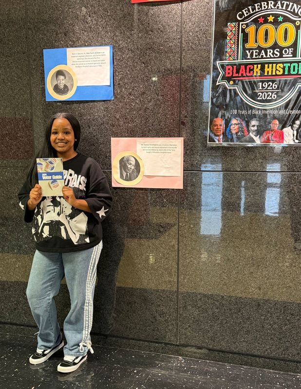 FBLA student standing in hallway with dark gray background holding up a sign with posters behind her on wall