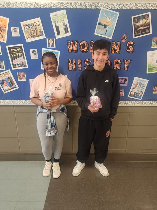 Two students stand in fron of a bulletin board holding their prizes for Fly High Fridays