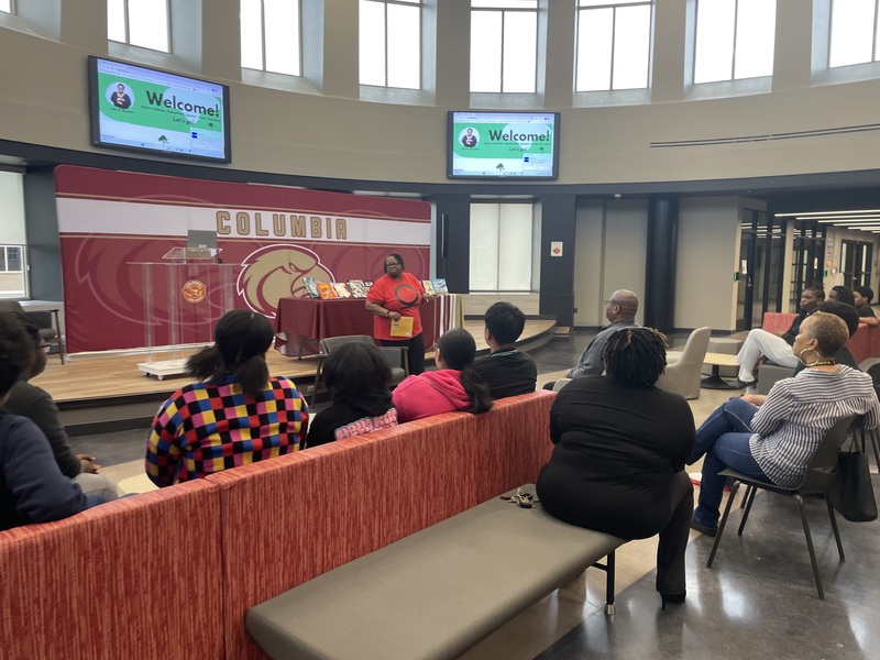 Dr. Royston, author holding up book  to english class with books she has written on display table behind her