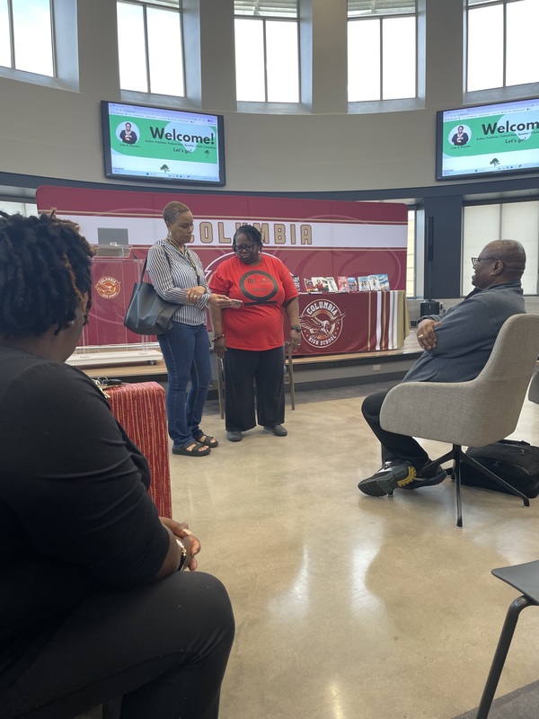 A parent standing talking to Dr. Royston showing her a book of illustrations while Mr. Royston and a friend sits in front of them