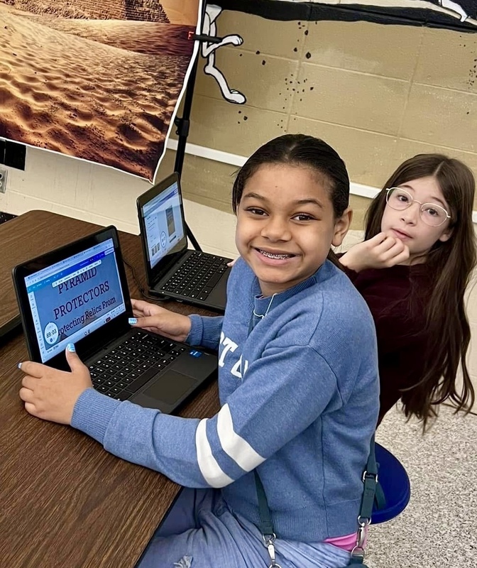 Two female students sitting at a desk with laptops smile as they look up into the camera