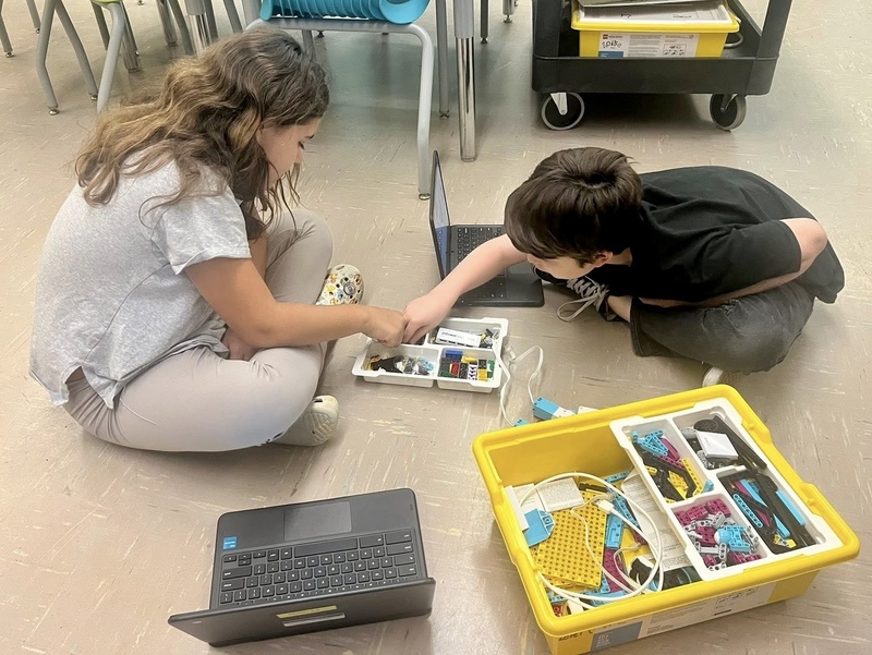 Two female students sitting on the floor reaching into a tray to collect robot parts