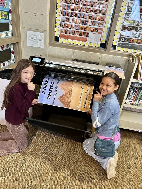 TWo girls working at a large printer smile back at the camera