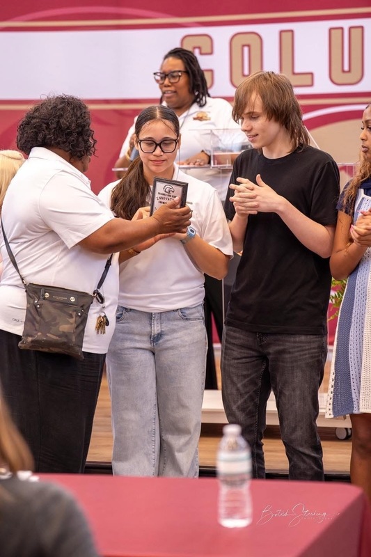 Mrs. Mastin, Assistant Principal handing trophies to two students while Dr. Brand stands in back on microphone.