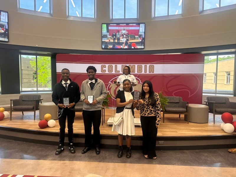 4 students standing in front of stage holding up plaques while Dr. Brand stands on the stage behind them with TV on wall showing the room