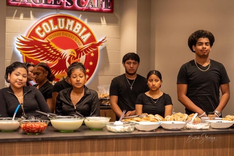 Six students from FACS class dressed in all black standing behind the counter of the Columbia Eagle's Cafe with sign on back wall