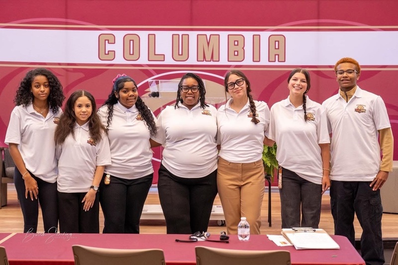 Dr. Brand, Curriculum Specilaist at Columbia standing in the middle of six students three on each side of her with Columbia Stand in the back of them
