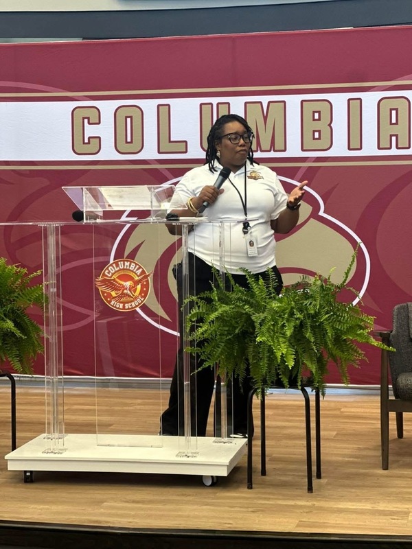 Dr. Brand standing on stage behind clear podium with two ferns on each side  and Columbia stand behind her