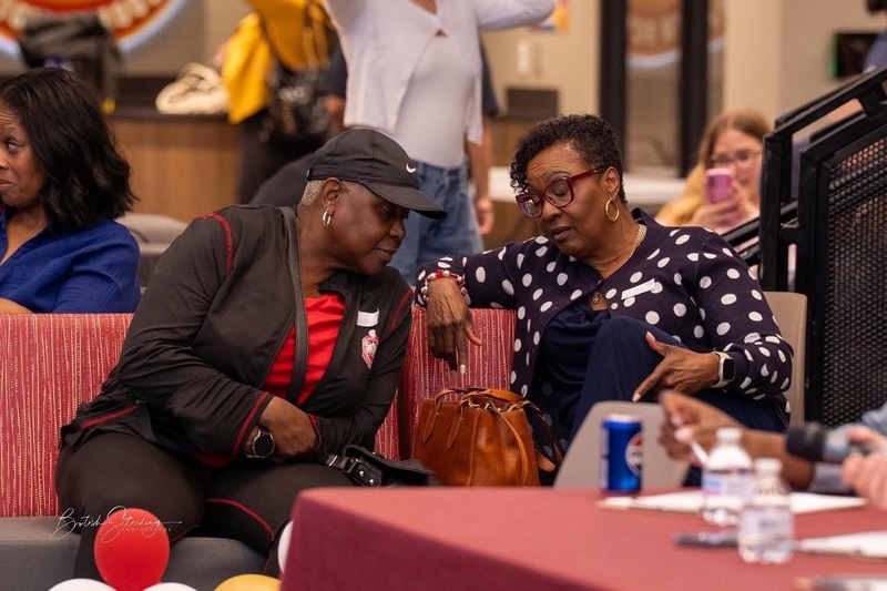 Two ladies on the couch with one leaning in to talk to the others with a red balloon in front