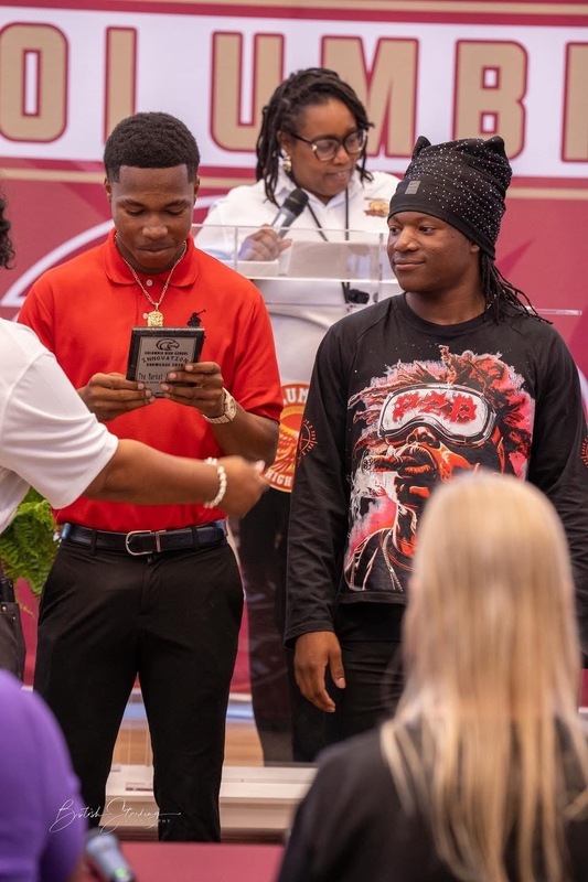 Two students standing in front of Dr. Brand on stage with one looking down at his trophy plague