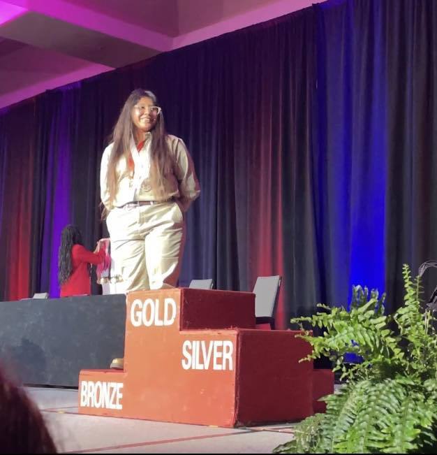 One student stands on a podium that says Gold, Silver, and Bronze. She is standing on the bronze side. 