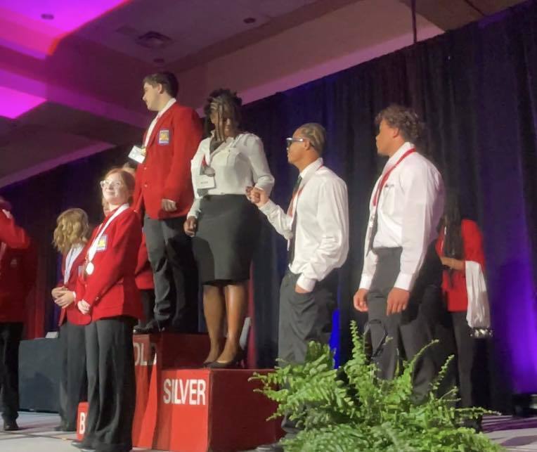 Multiple students stand on a podium that says Gold, Silver, and Bronze. Three of our students stand around the Silver side. Two students fist bump in celebration.