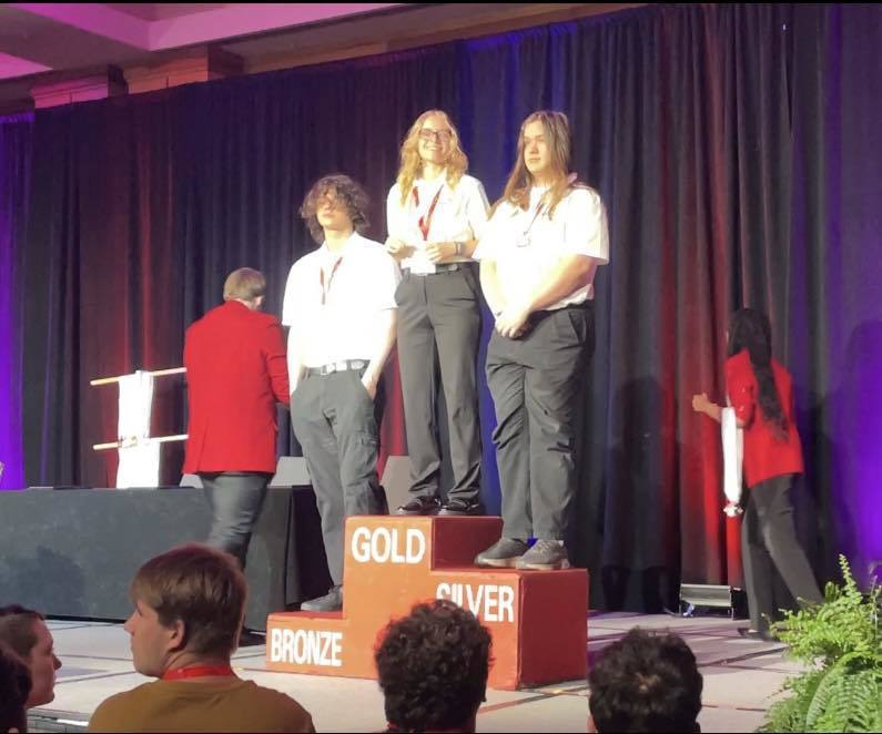 Three students stand on a podium that says Gold, Silver, and Bronze. Our student won Silver in this category.