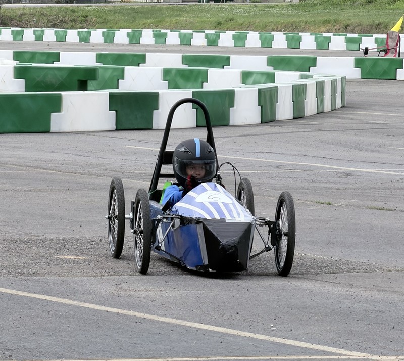 A Chapman Middle School student wearing a black racing helmet and blue racing shirt drives a blue and white GreenPower car around a paved track lined with green and white safety barriers.