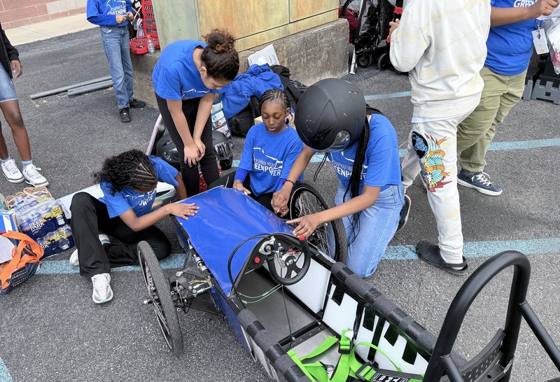 A group of students in blue "GreenPower" shirts work together in a parking lot to assemble the blue body panels of their custom-built electric race car.