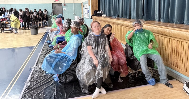 A picture of students in a long row of students and staff seated in chairs against a wooden stage. They are all wearing protective plastic ponchos and blue shoe covers, smiling and posing for the camera before the pie-throwing event begins.