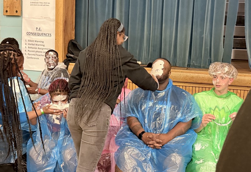 A student with long braids stands with her back to the camera, pressing a plate of whipped cream into the face of a seated staff member (Mr. Thornton). Other participants in ponchos sit nearby, already covered in whipped cream.