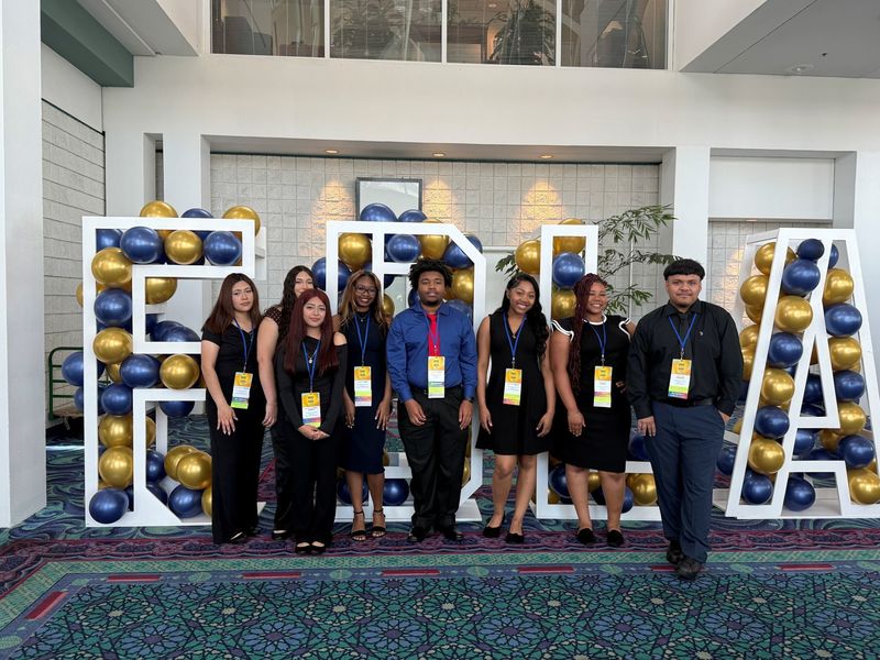 8 students of the Future Business Leaders of America dressed in dark blue and black posing for picture with Adviser standing in the middle with Large White Lettere, FBLA with blue and gold balloons