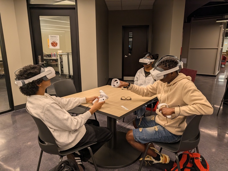 Three students sitting at table with VR headsets on with wands in hand during chemistry lesson