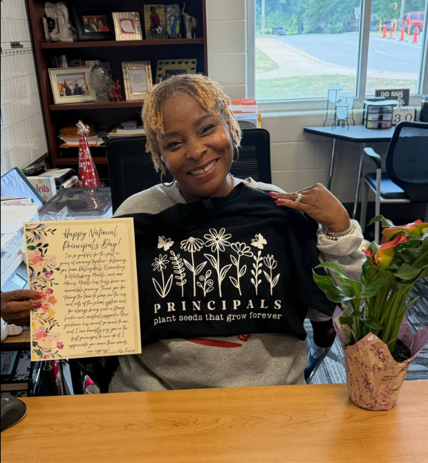 Dr. Burruss posing with a card and a tee shirt with flowers and the words "Principals plant seeds that grow forever".