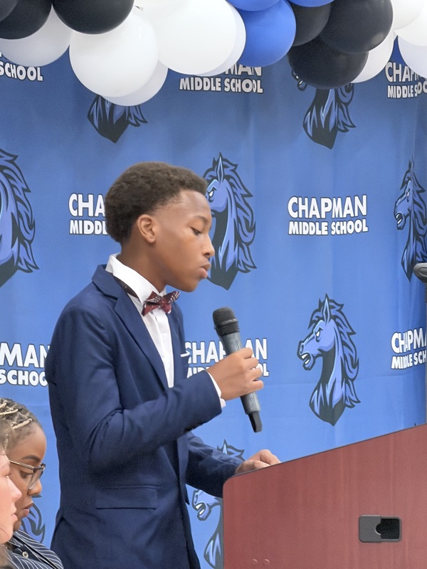 A young man wearing a navy blue suit and a patterned bow tie stands at a wooden podium, speaking into a microphone. Behind him is a blue backdrop featuring the Chapman Middle School logo and horse mascot, framed by a blue, black, and white balloon arch.