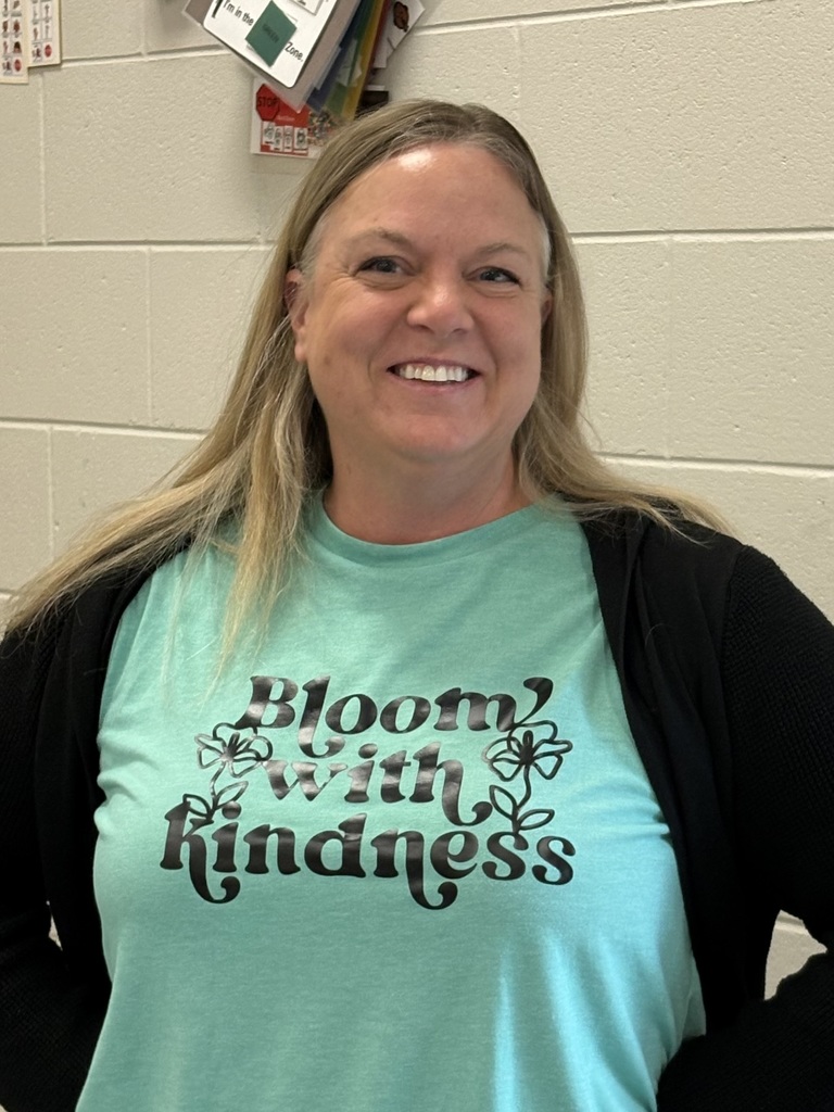  An educator standing against a wall wearing a blue shirt that reads “Bloom with Kindness.”
