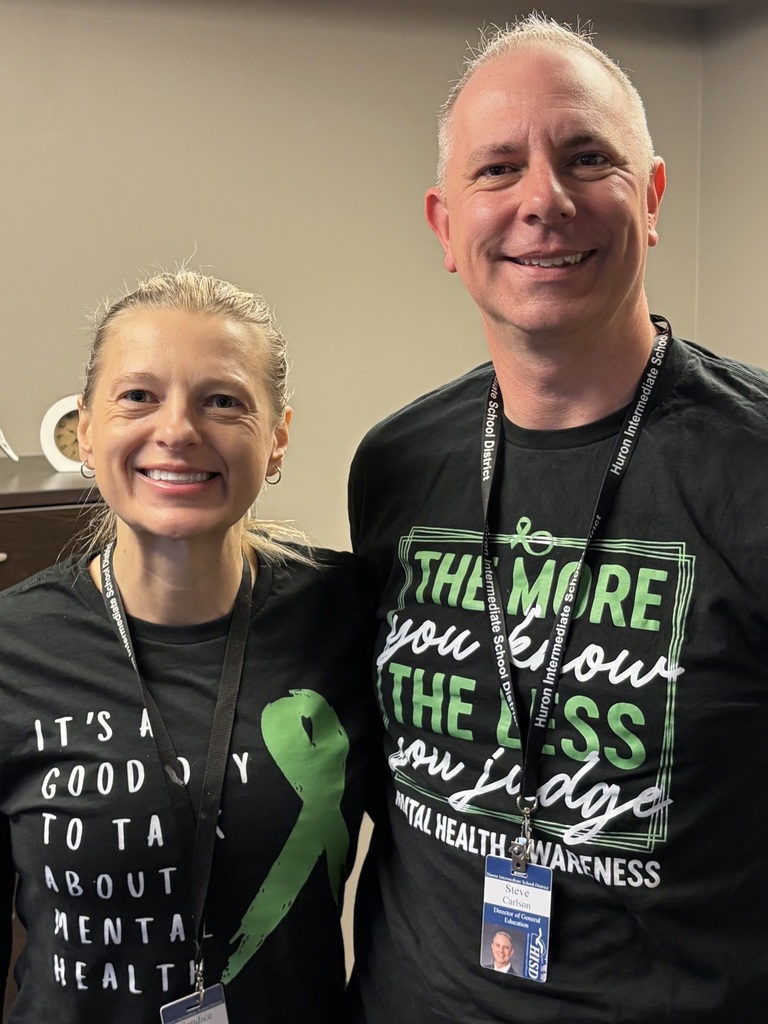Two educators standing close together, smiling and wearing black shirts with green mental health awareness messages.